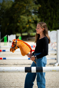 Lifestyle photo: a girl in a black top stands behind a jump pole on a sand arena holding the Hobby Horse Little Angel with Black Halter by Hobby Horse LarDen - chestnut plush head, bright red yarn mane - shown here with the Espresso fur halter in leatherette.