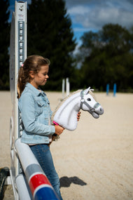 Lifestyle photo: a girl in a denim jacket stands by a jump wing on a sand arena holding the Hobby Horse Little Lady with Black Halter by Hobby Horse LarDen - white plush head, cream yarn mane - shown here with the grey halter with mint fur.