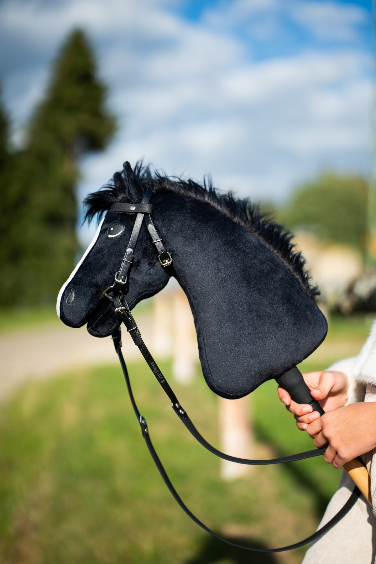 Detail photo: the head and neck of the Hobby Horse Loki with Black Halter by Hobby Horse LarDen held up in a meadow under a blue sky - black plush, white blaze, black yarn mane - shown here with the complete Pure Black bridle without noseband.