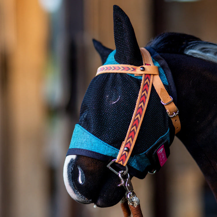 A close-up of a horse wearing a blue mesh fly mask with ear covers and a brown leather halter featuring a red arrow pattern, reminiscent of hobby horsing gear. The background is softly blurred.