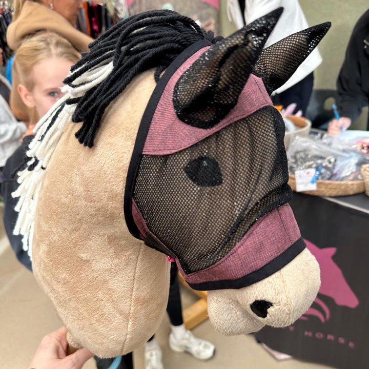 A close-up of a handmade Hobby Horse with a mesh mask and black-and-white yarn mane. In the blurred background, people and a booth with horse-themed items showcase the creative spirit of Hobby Horsing.
