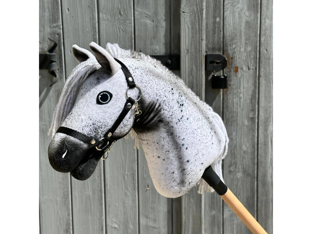 The Hobby Horse LarDen "Naivy" with a black halter is displayed in front of a rustic wooden door, showing off its gray and white Appaloosa styling and detailed mane.