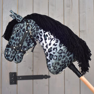 The Hobby Horse Picasso with Black Halter by Hobby Horse LarDen features a plush head with an Appaloosa-inspired gray and black spotted coat, a black halter, and a black yarn mane, all mounted on a wooden stick and shown against a gray wooden wall.