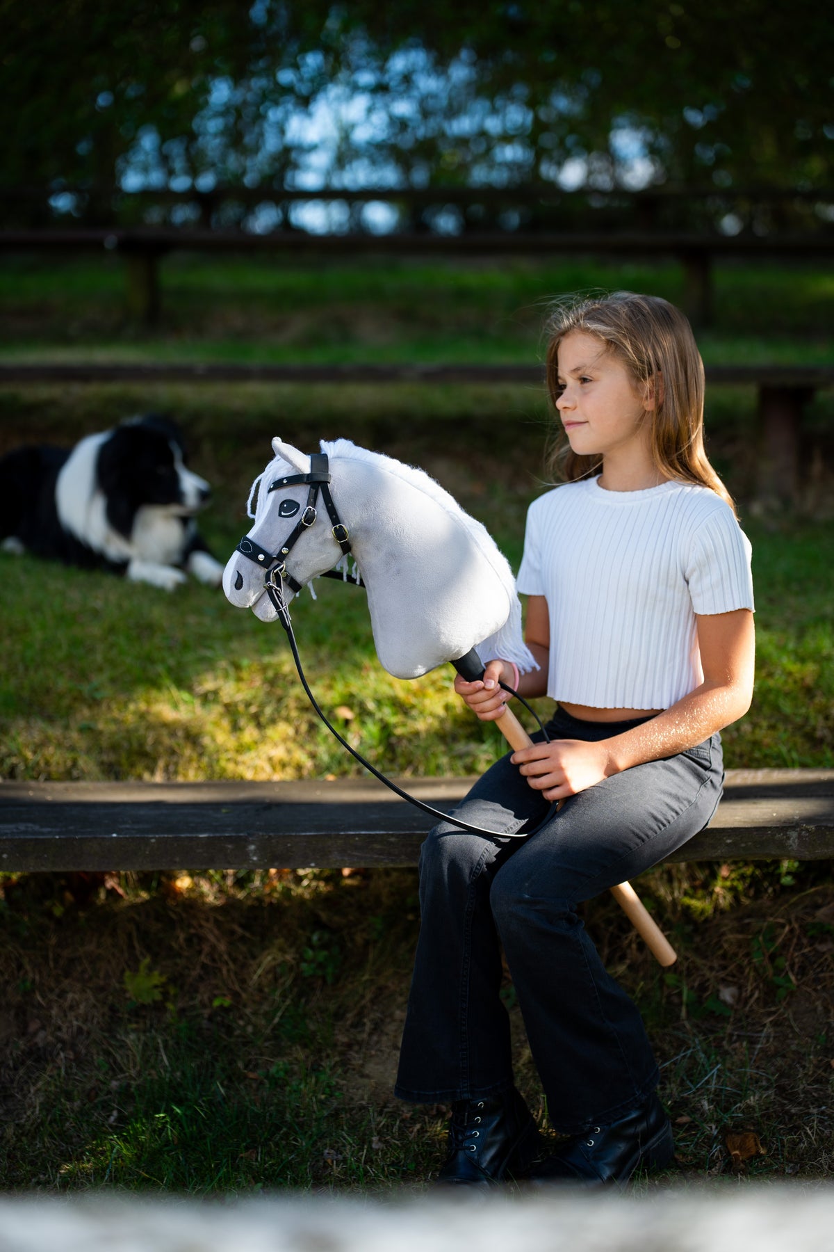 Lifestyle photo: a girl in a white top sits on a garden bench in the evening sun holding the Hobby Horse Popcorn with Black Halter by Hobby Horse LarDen, a border collie resting behind - white plush head, white yarn mane - shown here with the decorated bitless bridle.