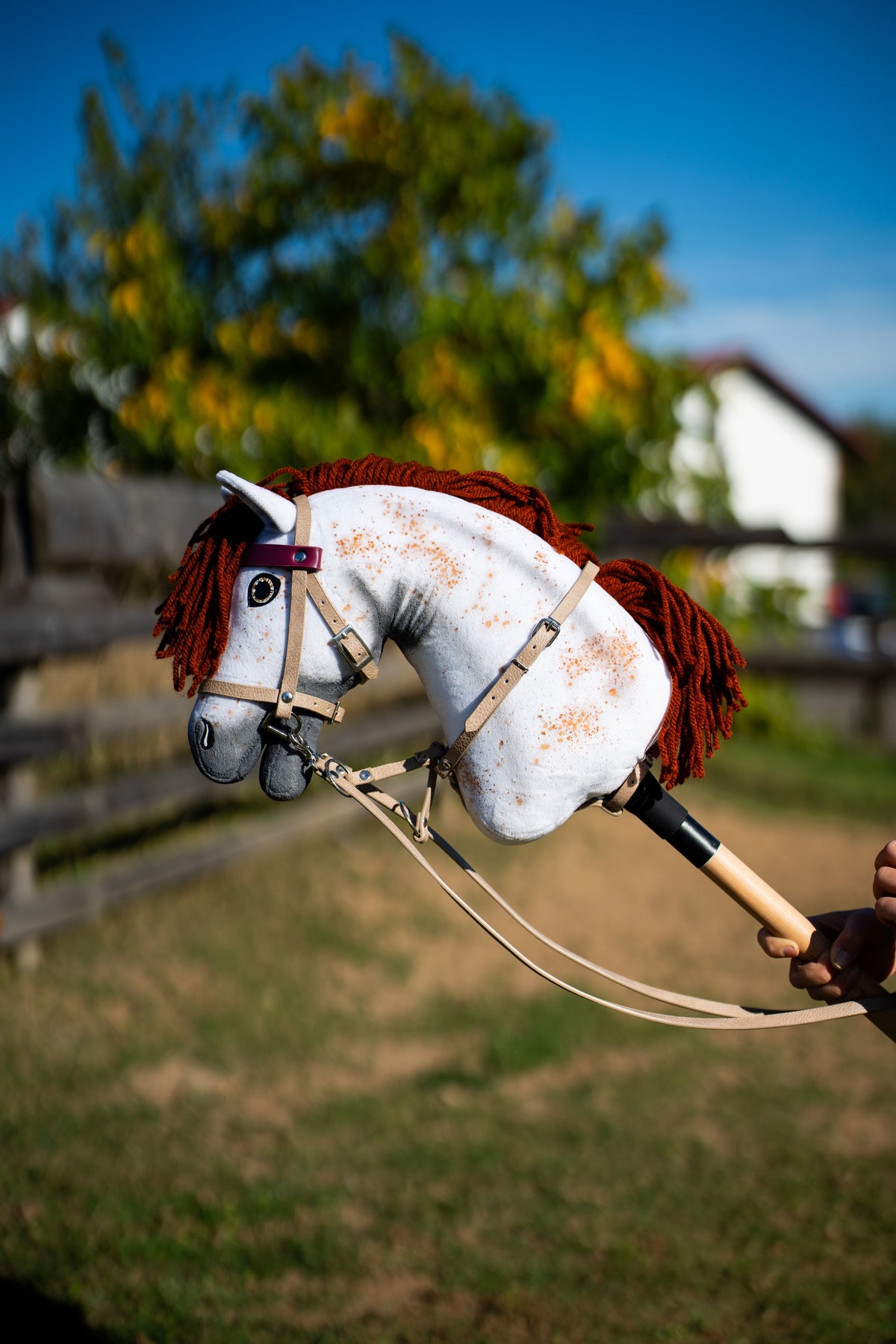 White speckled Hobby Horse with a red-brown yarn mane in the beige bridle and reins with bordeaux browband by a paddock fence