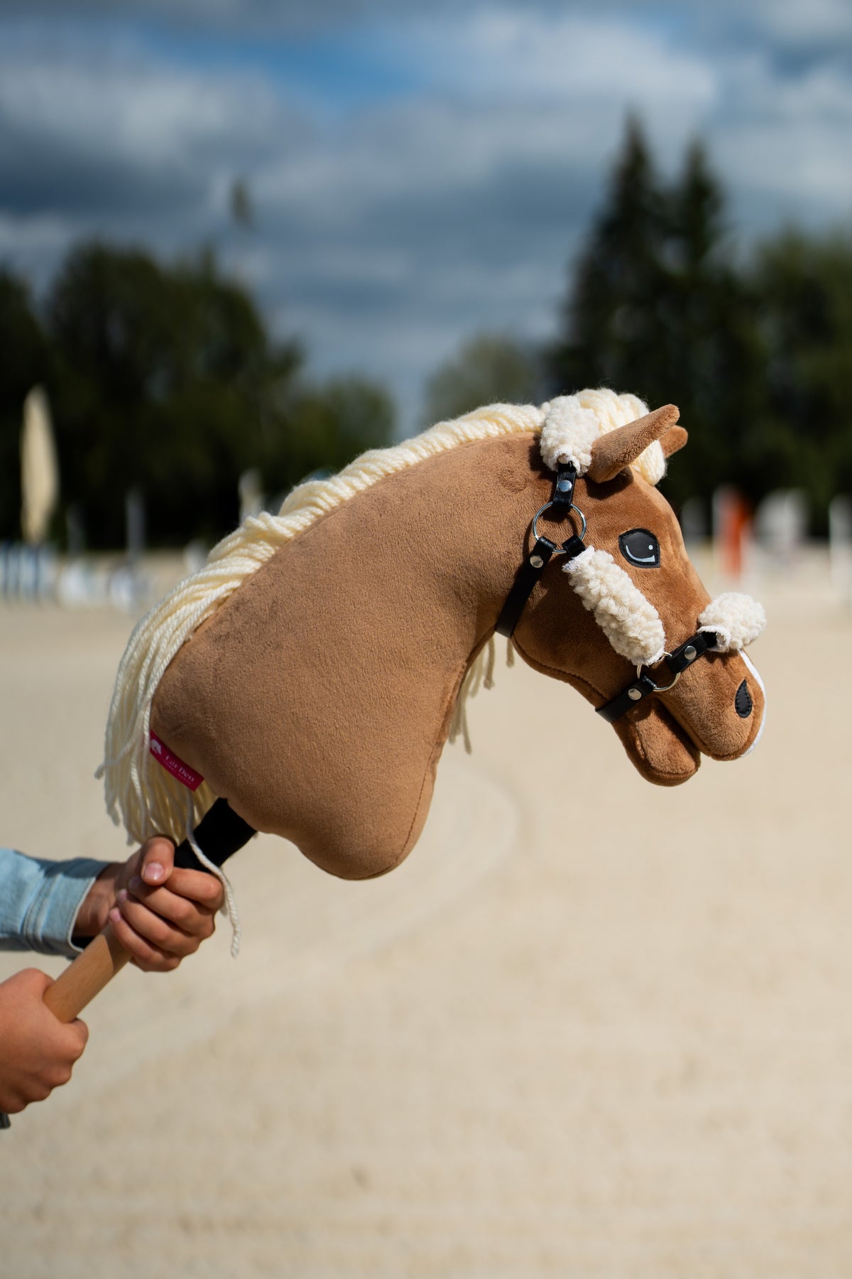Detail photo: the head of the Hobby Horse Smart with Black Halter by Hobby Horse LarDen held up on a sand arena under dark clouds - palomino plush, cream yarn mane, white fur noseband - shown here with the black leatherette halter with complete fur.