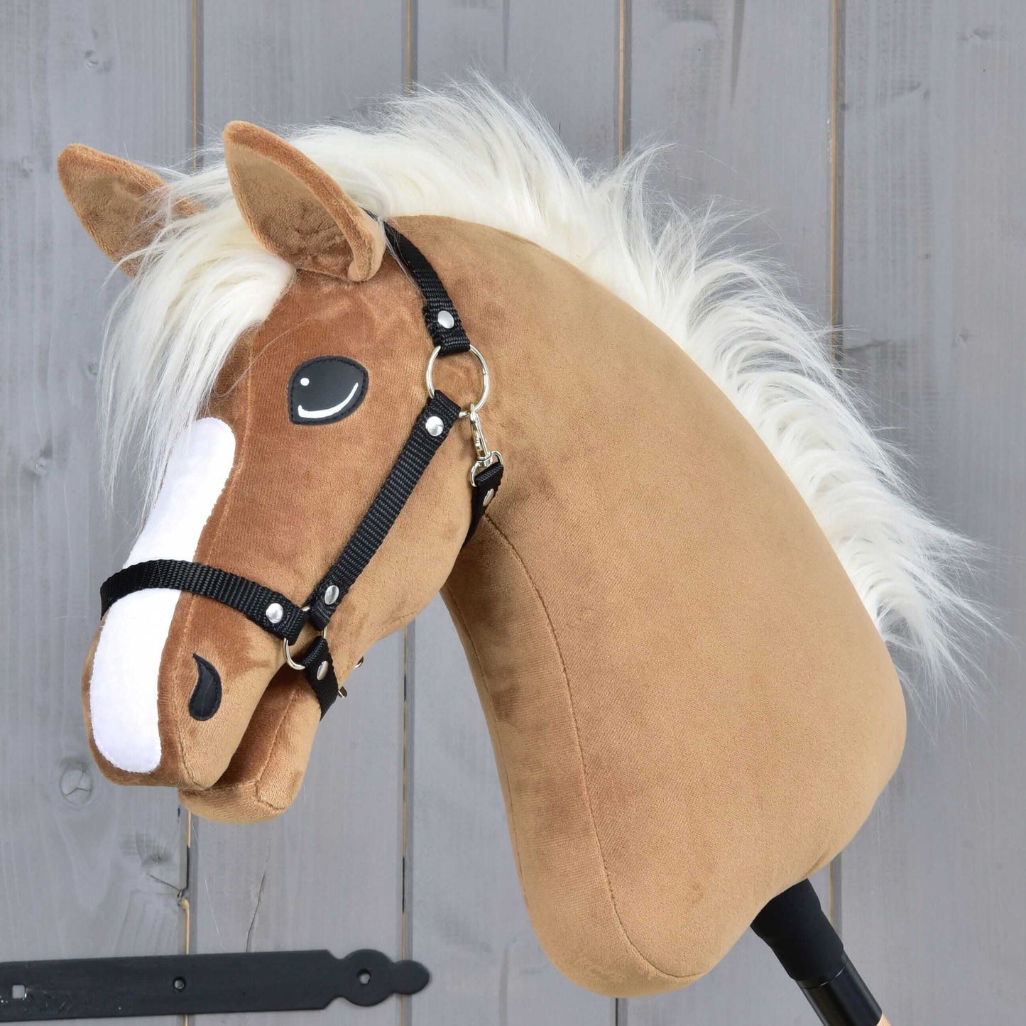 The Hobby Horse LarDen "Hobby Horse Star" with a black halter and a caramel coat, white mane, and black bridle stands against a gray wooden background.