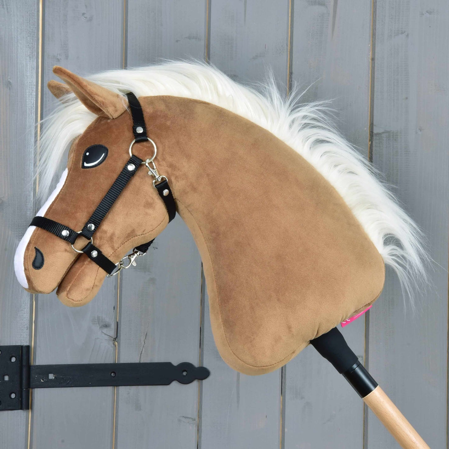 The Hobby Horse LarDen "Hobby Horse Star with black halter" features a brown plush head with a caramel coat, white mane, black halter, and white nose markings on a wooden stick in front of a gray wooden plank wall.