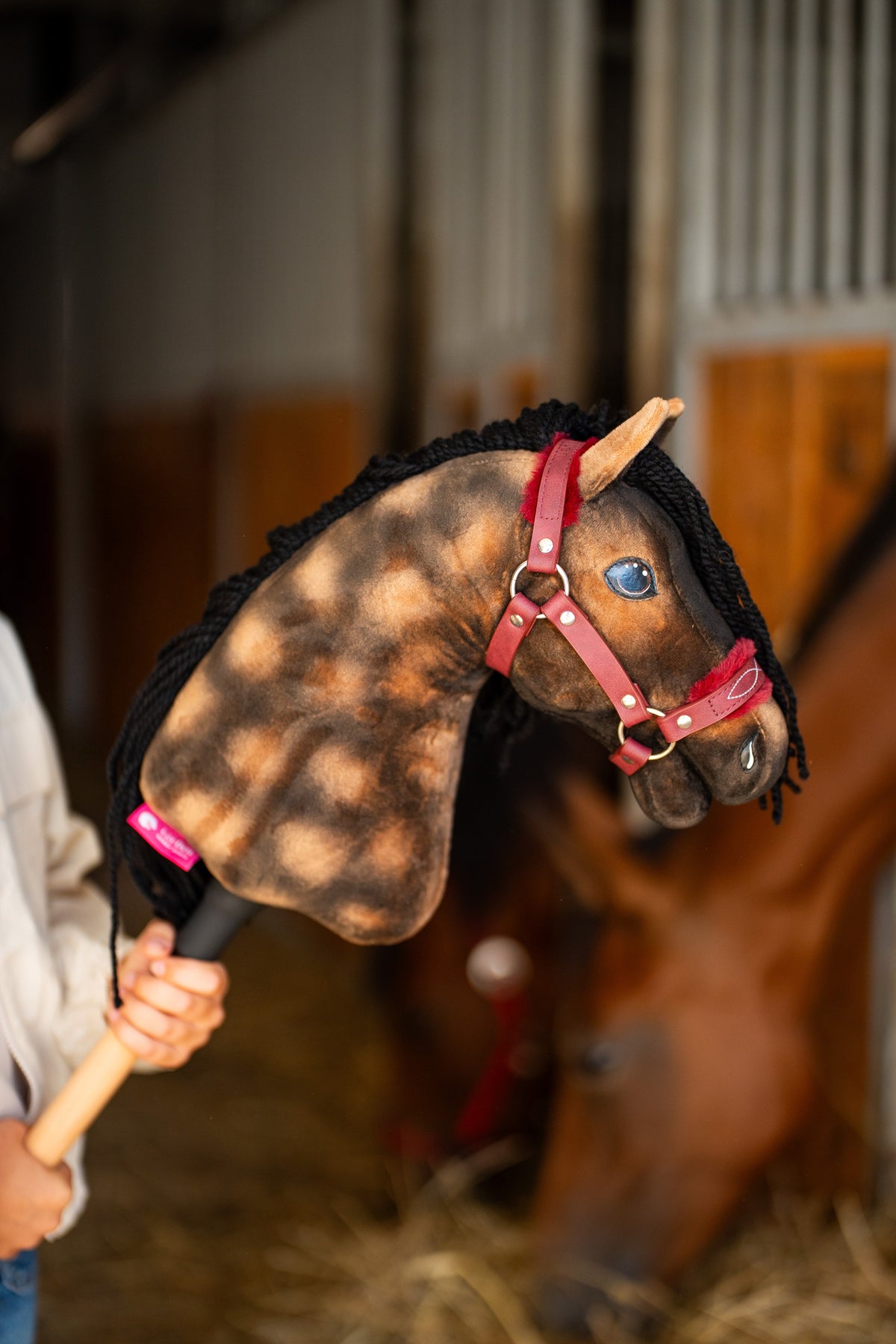 Detail photo: the head of the Hobby Horse Stormy with Black Halter by Hobby Horse LarDen held up in a stable, a real bay horse blurred behind - dappled brown plush, blue eye, black yarn mane - shown here with the Burgundy fur halter in leatherette.