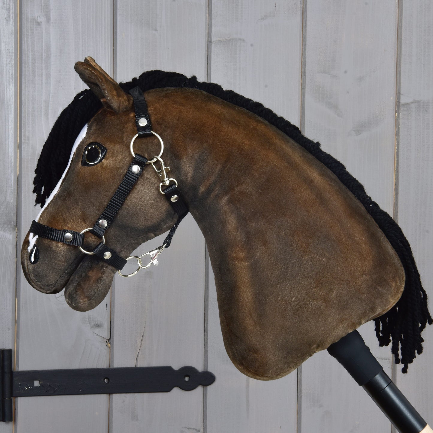 The Hobby Horse LarDen "Hobby Horse Sundance with Black Halter" features a plush design with a black yarn mane, bridle, and reins—capturing the spirited look of an English Thoroughbred stallion—and is mounted on a stick, shown against a light wooden plank wall.