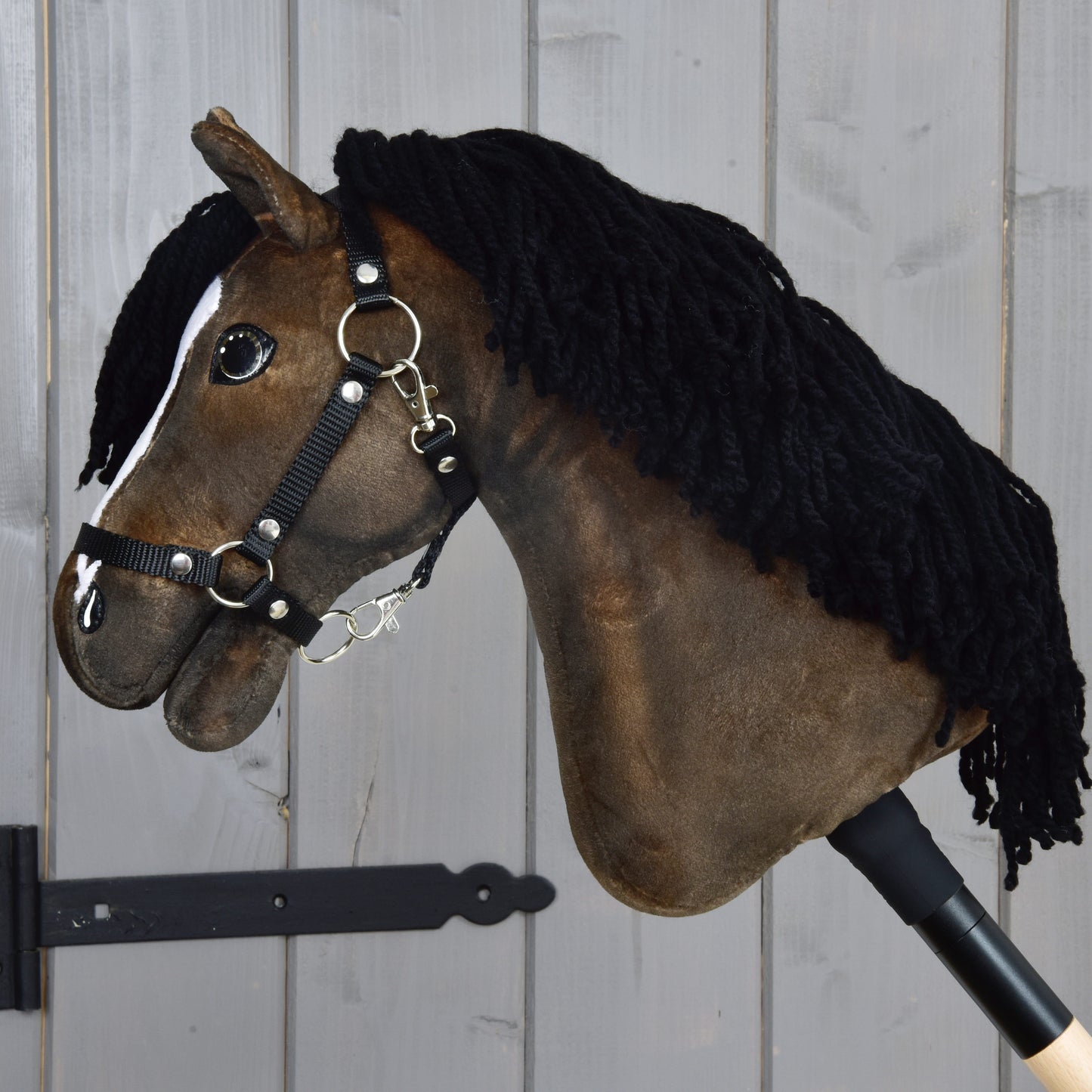 The Hobby Horse LarDen "Hobby Horse Sundance with Black Halter" is a brown fabric hobby horse inspired by the swift English Thoroughbred breed, featuring a thick black yarn mane, a white blaze on its face, and a black nylon halter with silver buckles. It is mounted on a stick and displayed against a grey wooden background.