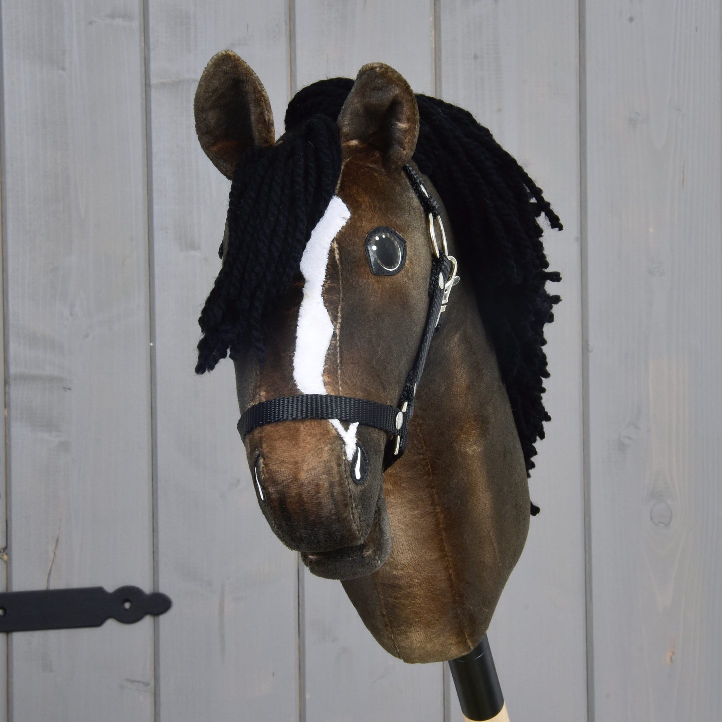 Hobby Horse LarDen’s Hobby Horse Sundance with Black Halter features a brown coat, black yarn mane, striking white blaze, and black halter—styled after an English Thoroughbred stallion. Set against a light wooden wall, this charming creation celebrates one of the fastest horse breeds.