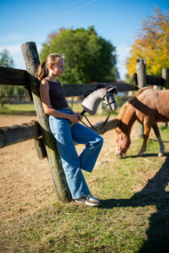 Lifestyle photo: a girl in a brown top and jeans leans on a wooden fence post holding the Hobby Horse Thor with Black Halter by Hobby Horse LarDen - grey plush head with a black yarn mane - a pony grazing in the paddock behind her, shown here with the Complete black bridle with diamonds.